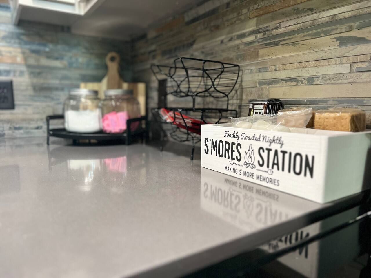 A kitchen counter with a sign for S'mores Station, jars of marshmallows, and a basket of graham crackers.