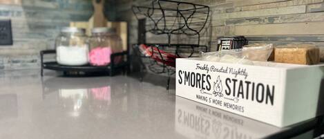 A kitchen counter with a sign for S'mores Station, jars of marshmallows, and a basket of graham crackers.