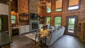 A cozy cabin interior with a stone fireplace, wooden walls, a sofa, and a dining area. - 3 BR Luxury Cabin & Hot Tub near Buffalo River! (Jasper)