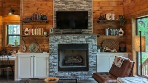 A cozy log cabin interior with a stone fireplace, a comfortable seating area, and a television mounted above the fireplace. - 3 BR Luxury Cabin & Hot Tub near Buffalo River! (Jasper)