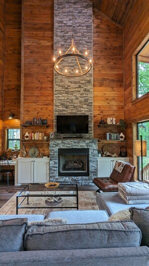 L’intérieur chaleureux d’un chalet en bois, avec une cheminée en pierre, un coin salon confortable et un téléviseur fixé au-dessus de la cheminée.