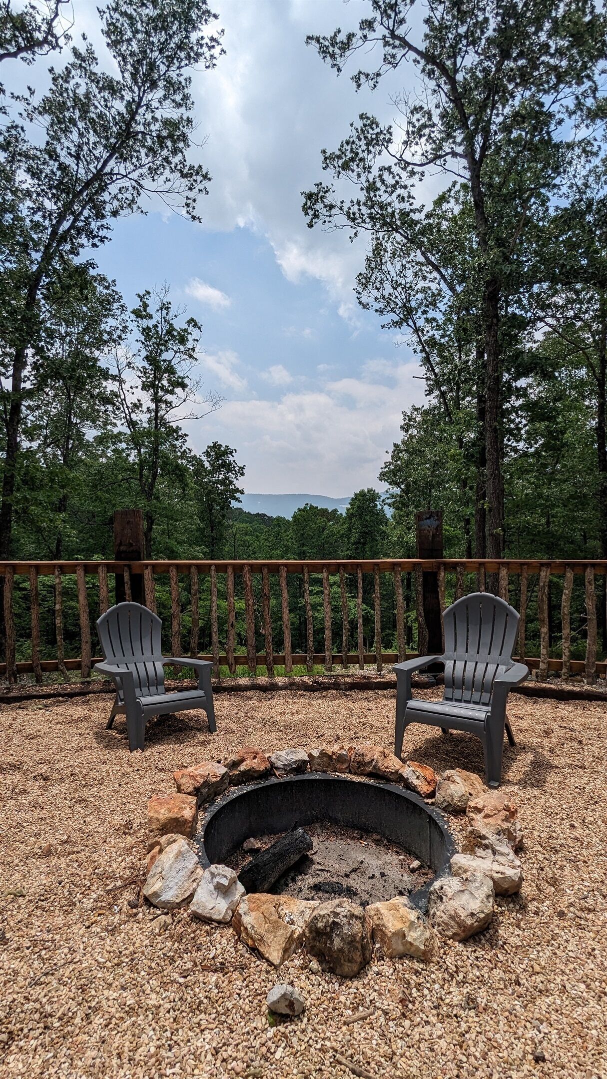 An outdoor seating area with two Adirondack chairs, a fire pit, and a wooden deck overlooking a forested landscape.