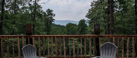 An outdoor seating area with two Adirondack chairs, a fire pit, and a wooden deck overlooking a forested landscape.