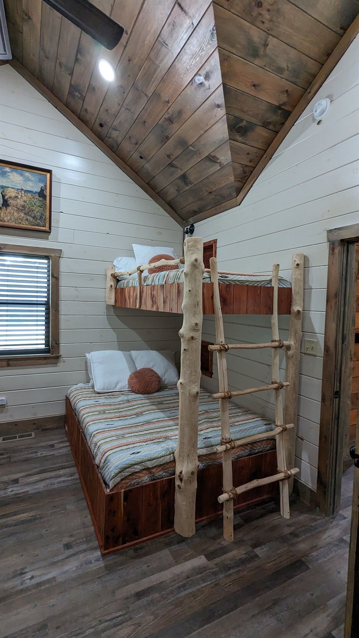 A bunk bed with a ladder, wooden walls, a window with blinds, and a ceiling fan.