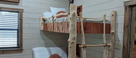 A bunk bed with a ladder, wooden walls, a window with blinds, and a ceiling fan.