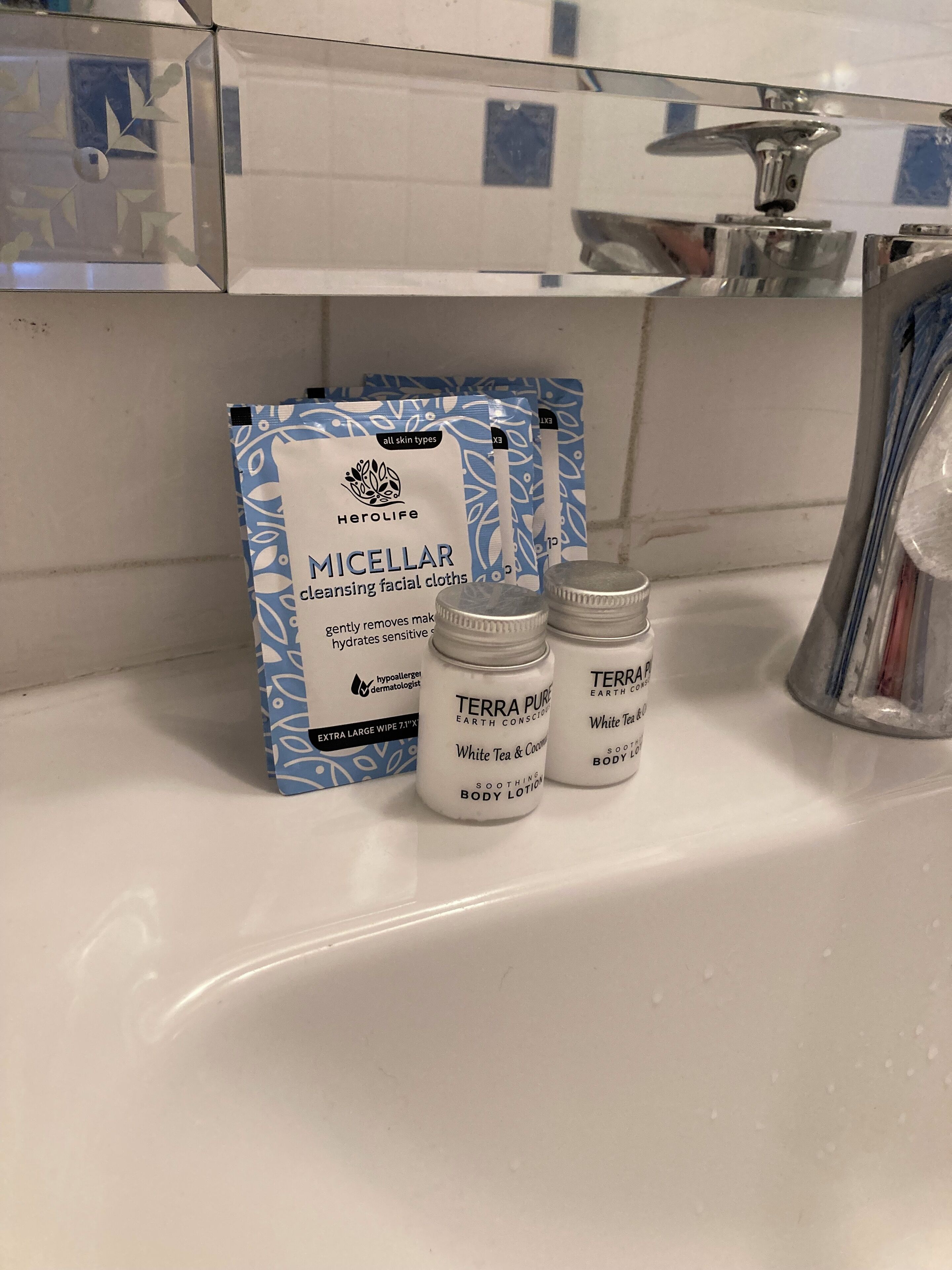 A bathroom sink with a white countertop. On the counter, there are two small white jars labeled 'Terra Pure Earth Concoction' and 'White Tea & Coconut Body Lotion'. Next to them is a blue and white package labeled 'Herolife Micellar Cleansing Facial Cloths'.