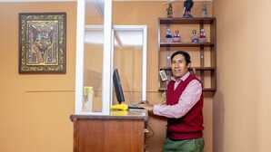 A man at a desk with a computer, a framed artwork on the wall, and a shelf with decorative items. - Colibri Inn (Chivay)
