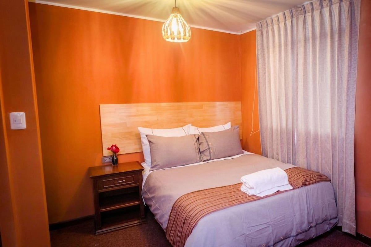Una habitación de hotel con una cama, una mesita de noche, una lámpara y un ventanal con cortinas.