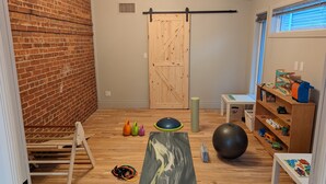 Una stanza con parete in mattoni, porta in legno, una sbarra per trazioni, un tappetino da yoga, un ruotolo da yoga, un balón medicinale e uno scaffale con giochi e libri.