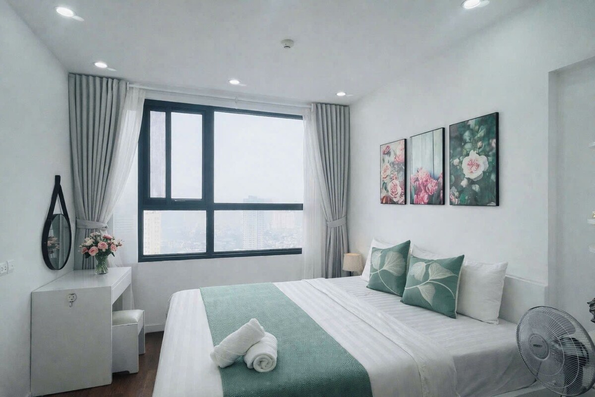 A hotel room with a bed, a desk, a chair, a standing fan, and a window with curtains.