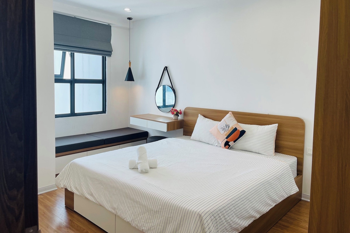 A modern bedroom with a large bed, a bedside table, a bench, a window with blinds, a hanging mirror, and a ceiling light.