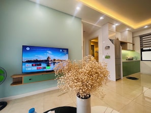 A modern living room with a flat-screen TV, a decorative vase with dried flowers, and a green kitchen area in the background. - Luna's House in Vinhomes D'Capitale (Hanoi)