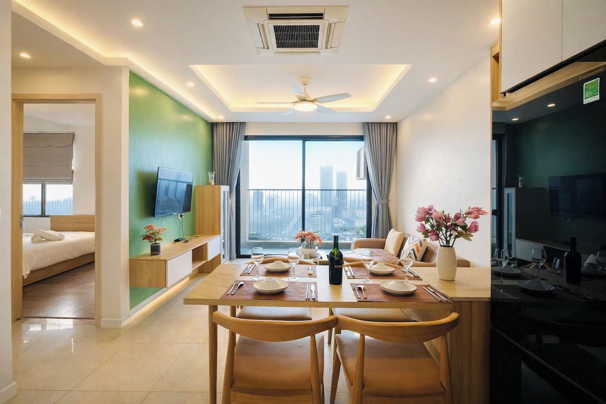 A modern hotel room with a dining area, a TV, a balcony with a city view, and a kitchenette.