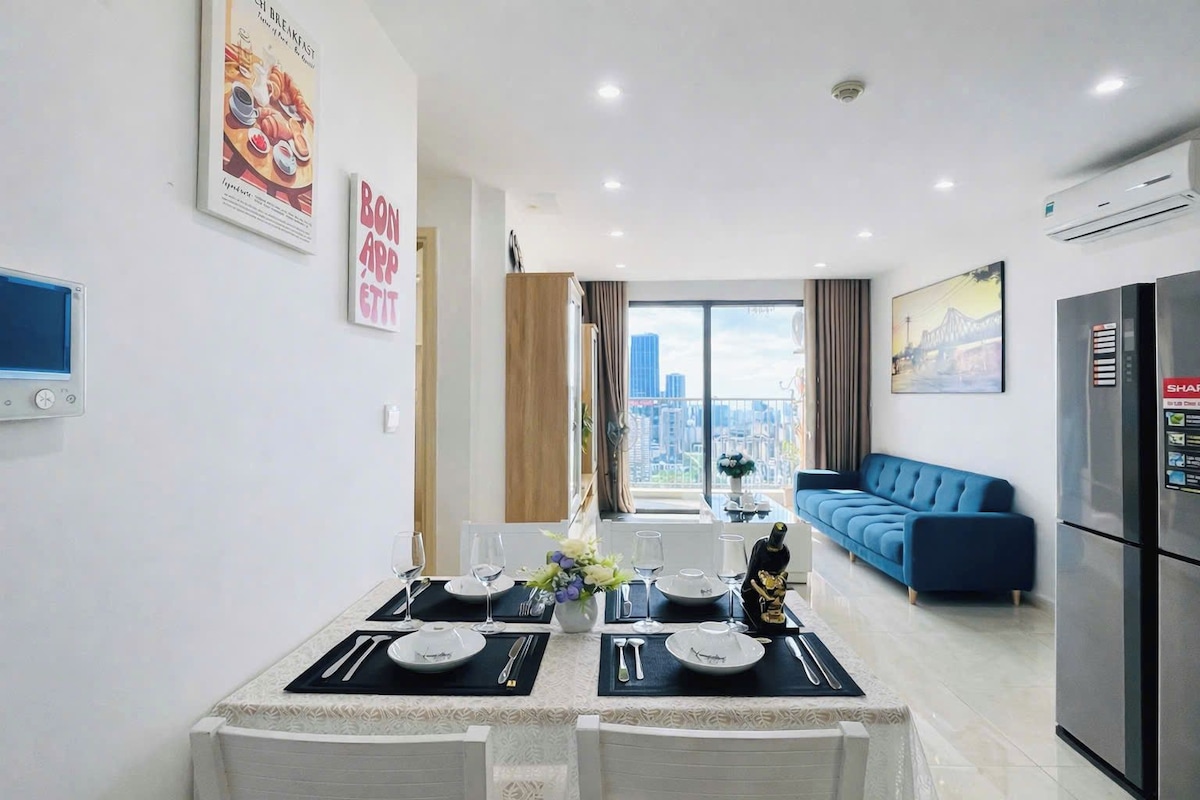 A modern living space with a dining area set for two, a blue sofa, and a view of the cityscape.