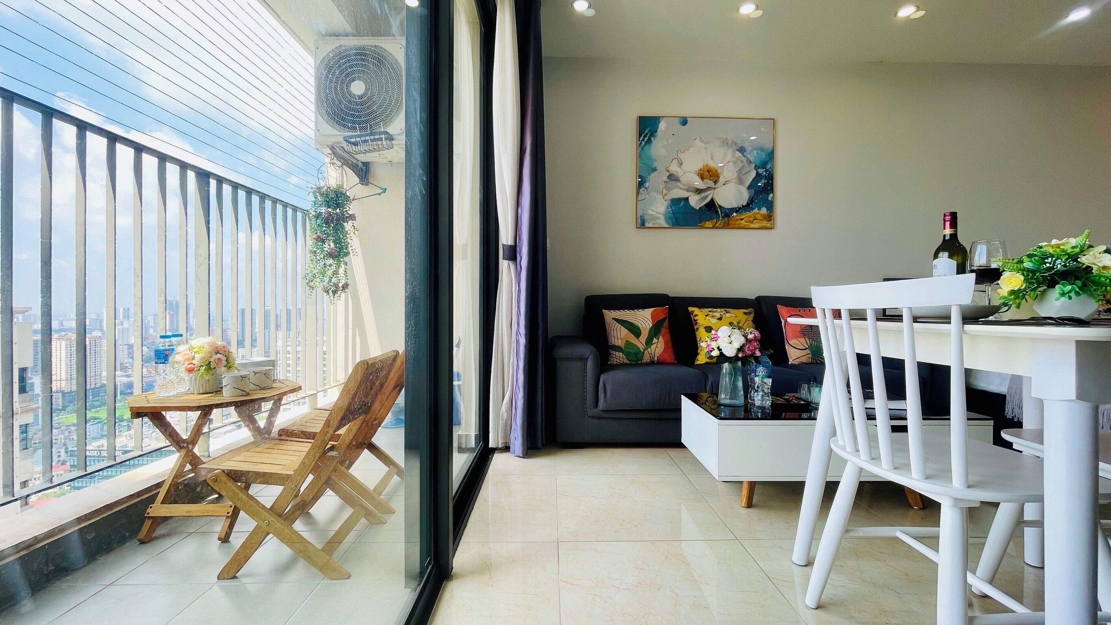 A modern living room with a dining area, a sofa with colorful cushions, a painting on the wall, and a balcony with a view of the city.