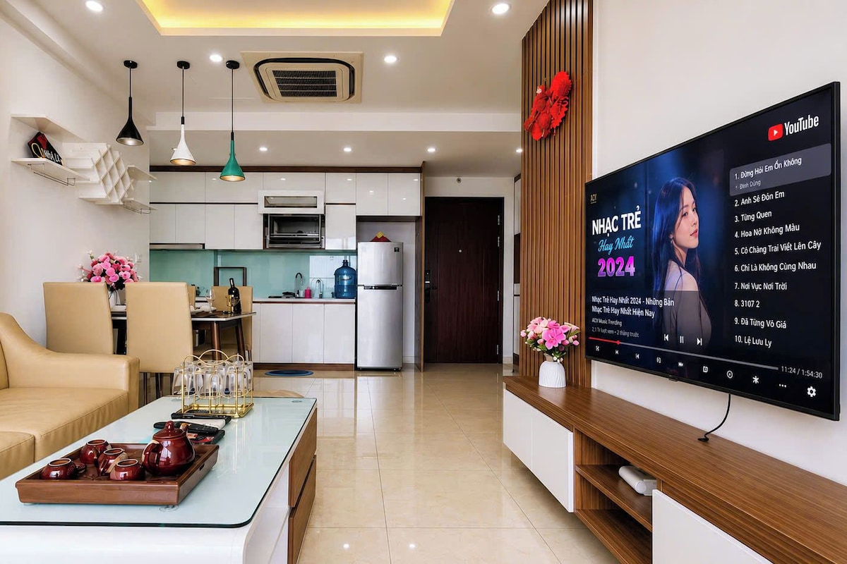 A modern living room with a sofa, coffee table, and a flat-screen TV displaying a music video.