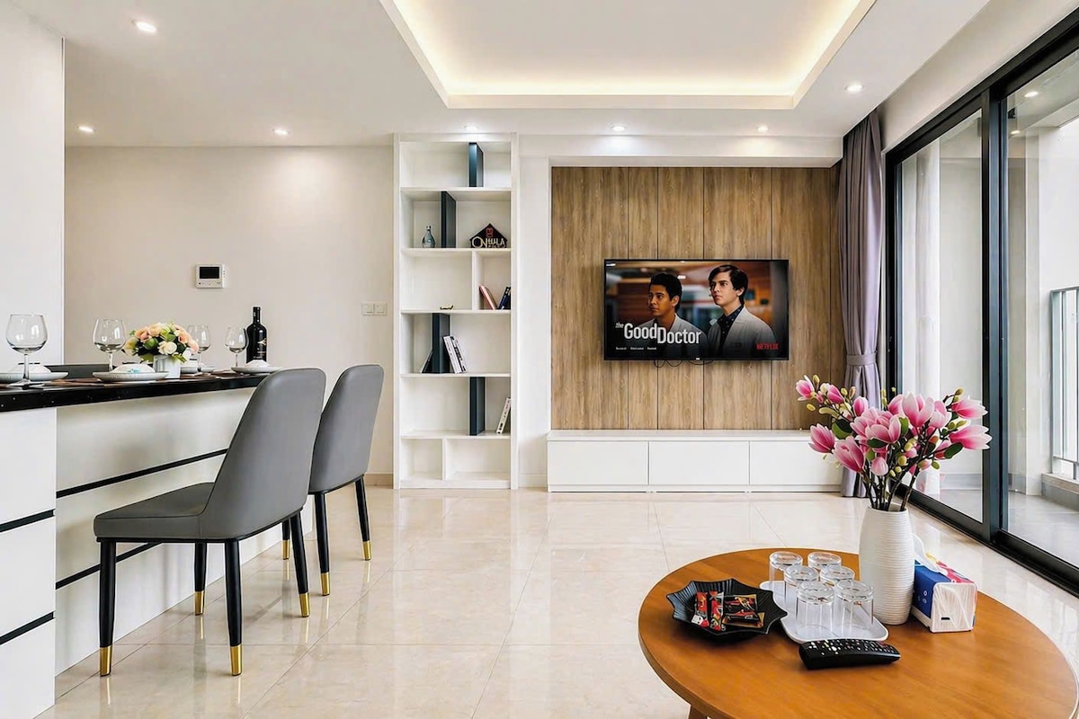 A modern hotel room with a dining area, a TV mounted on a wooden wall, and a large window offering a city view.