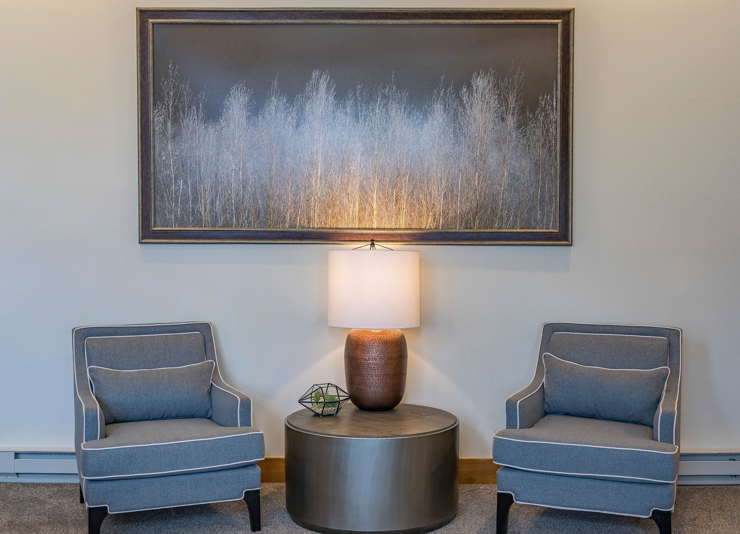 Una habitación con dos sillones grises, una mesita redonda de metal con una lámpara, y un cuadro abstracto grande en la pared.