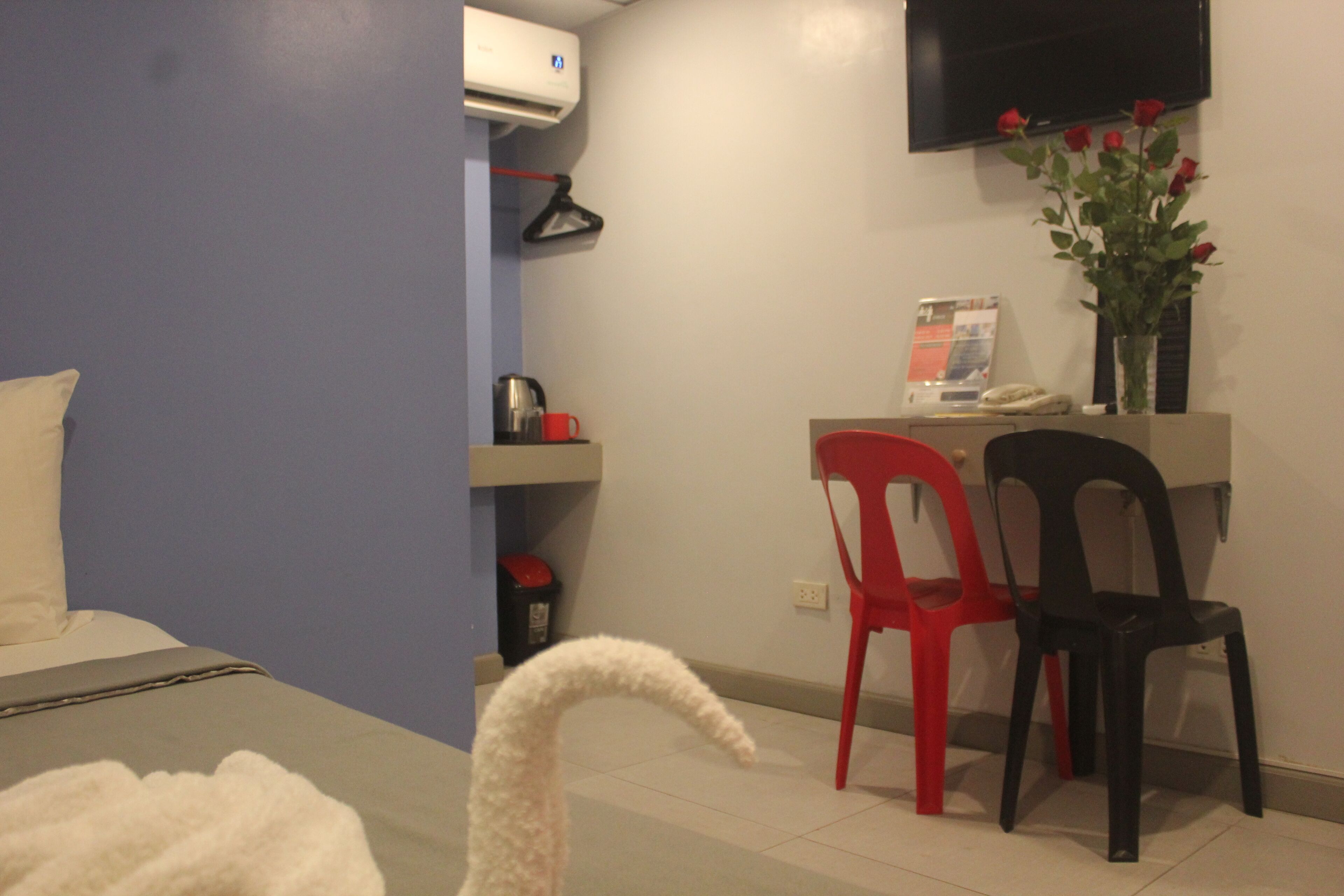 A hotel room with a bed, a desk with a chair, a TV, a vase with flowers, and a red and black chair.