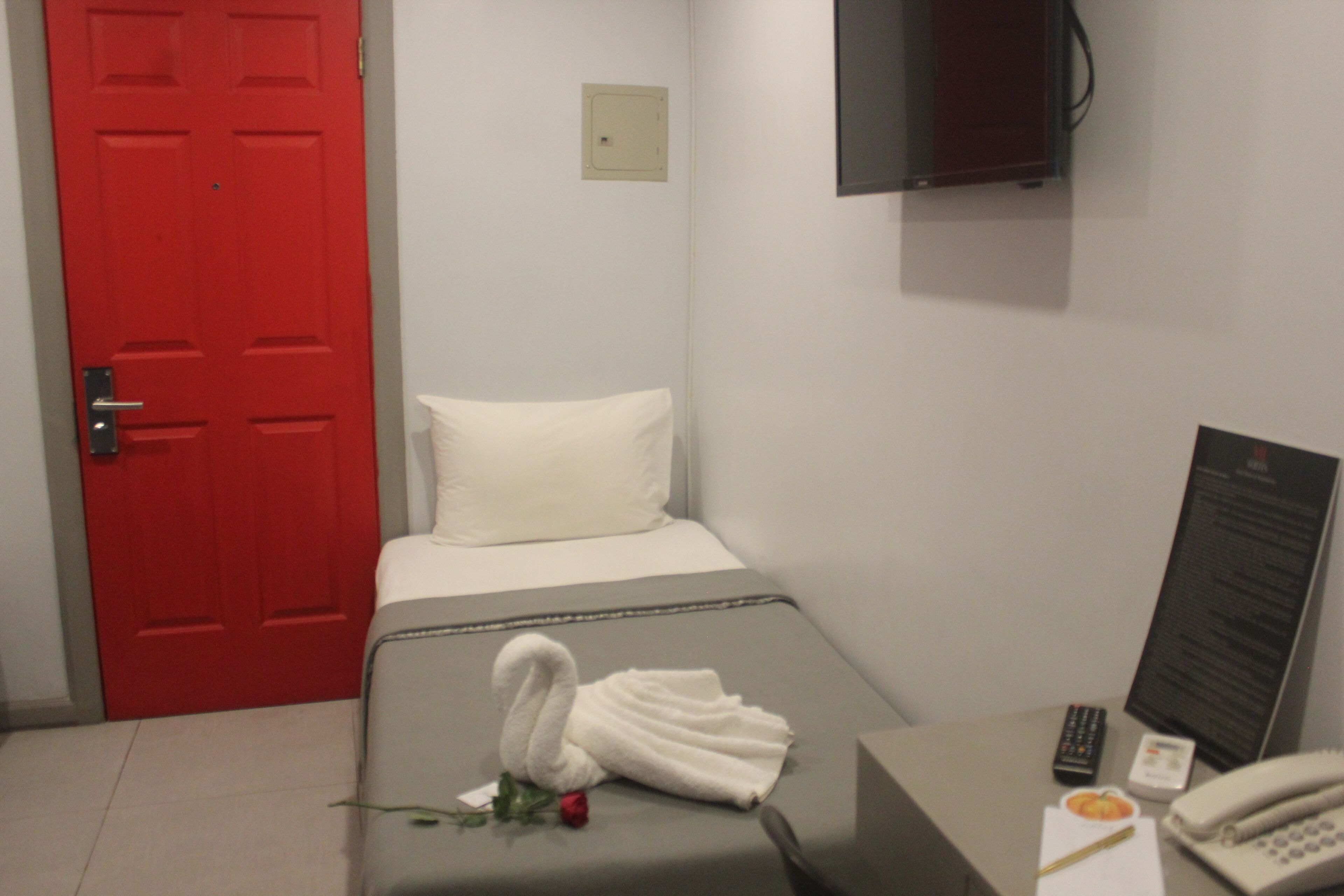 A hotel room with a red door, a bed with white bedding, a small table with a telephone, a remote control, and a menu board.