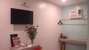 Une pièce avec un téléviseur à écran plat fixé au mur, un vase avec des fleurs rouges posé sur une table, une chaise rouge, un extincteur et une étagère sur laquelle se trouve un petit objet.