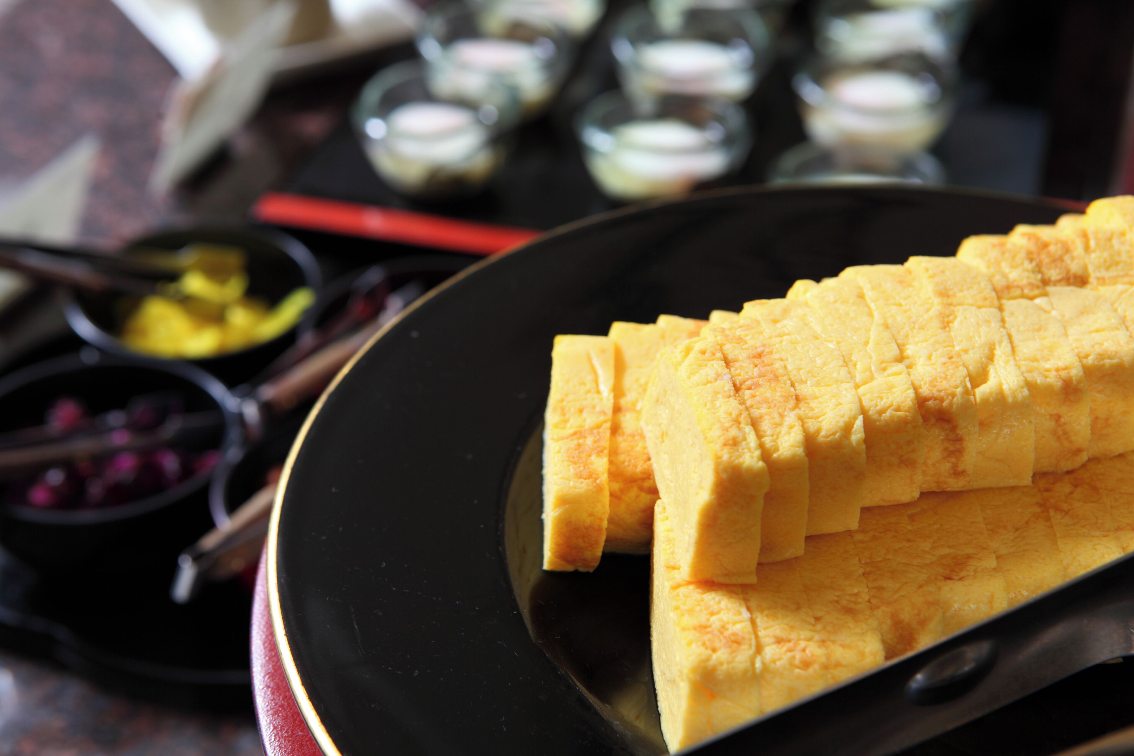 Une assiette noire sur laquelle sont disposés des aliments jaunes, coupés avec soin, peut-être des fruits ou des légumes, sur une table. En arrière-plan, on aperçoit un objet rouge et de petits bols.