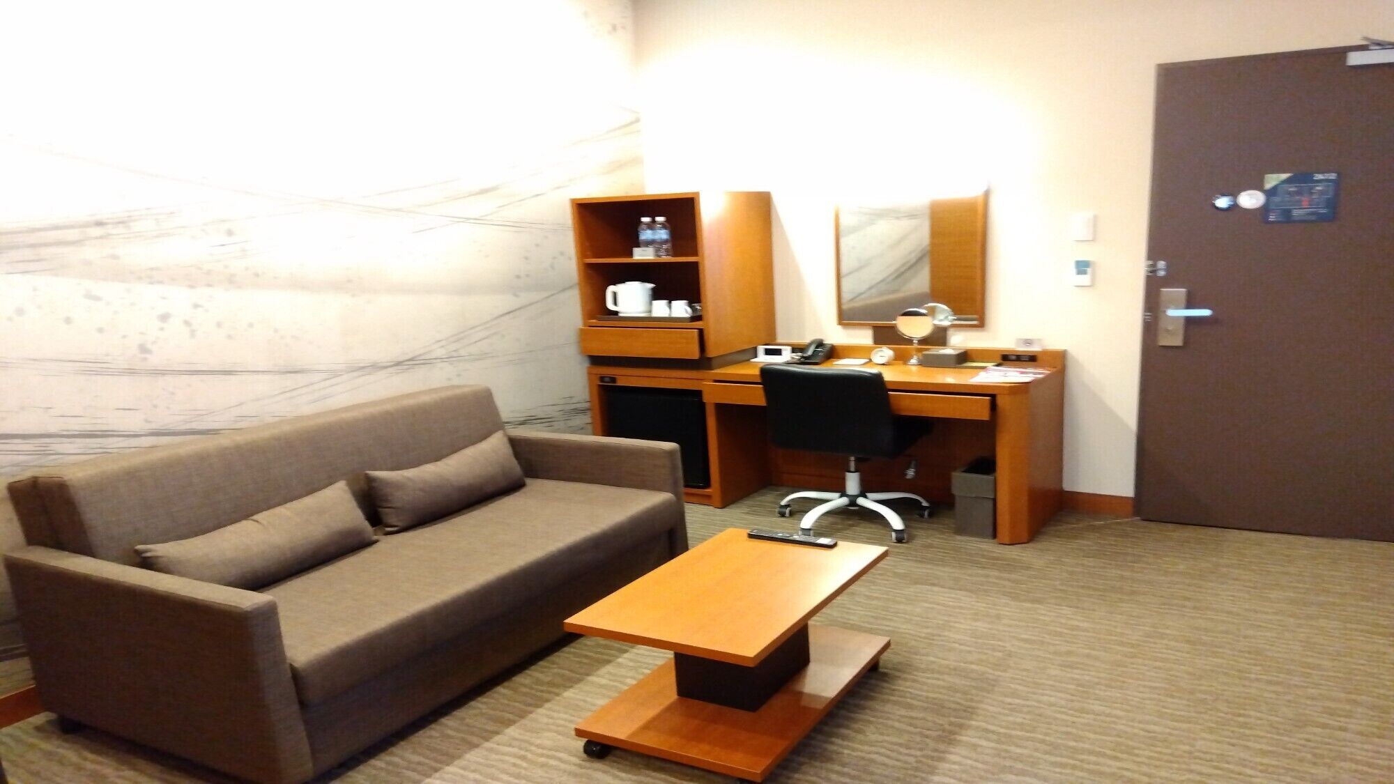 A hotel room with a sofa, a desk with a chair, a small table, a mirror, a shelf with toiletries, and a door.