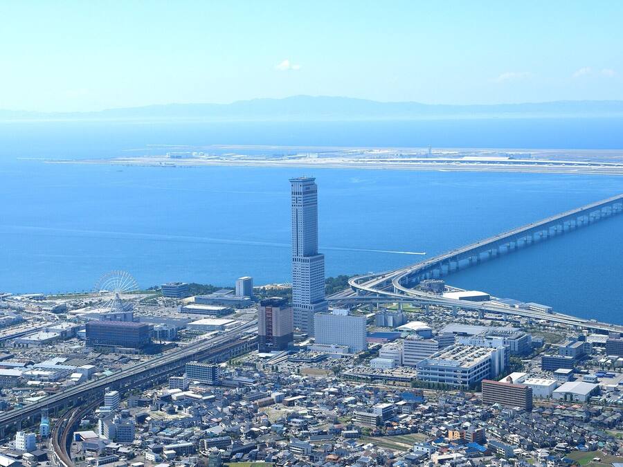 An aerial view of a coastal city with a prominent skyscraper, a long bridge, and a dense urban area with various buildings.