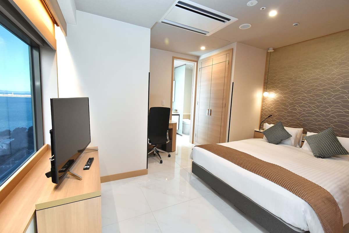 A modern hotel room with a large bed, a desk with a computer, a TV, and a view of the sea.