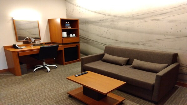 A hotel room with a wooden desk, a black office chair, a brown sofa, a small wooden coffee table, and a minibar with bottled water and tea. - Odysis Suites Osaka Airport Hotel (Izumisano)