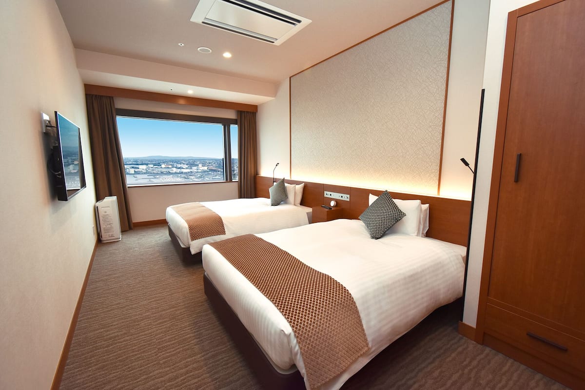 A hotel room with two beds, a large window with a city view, a flat-screen TV, and a wooden door.