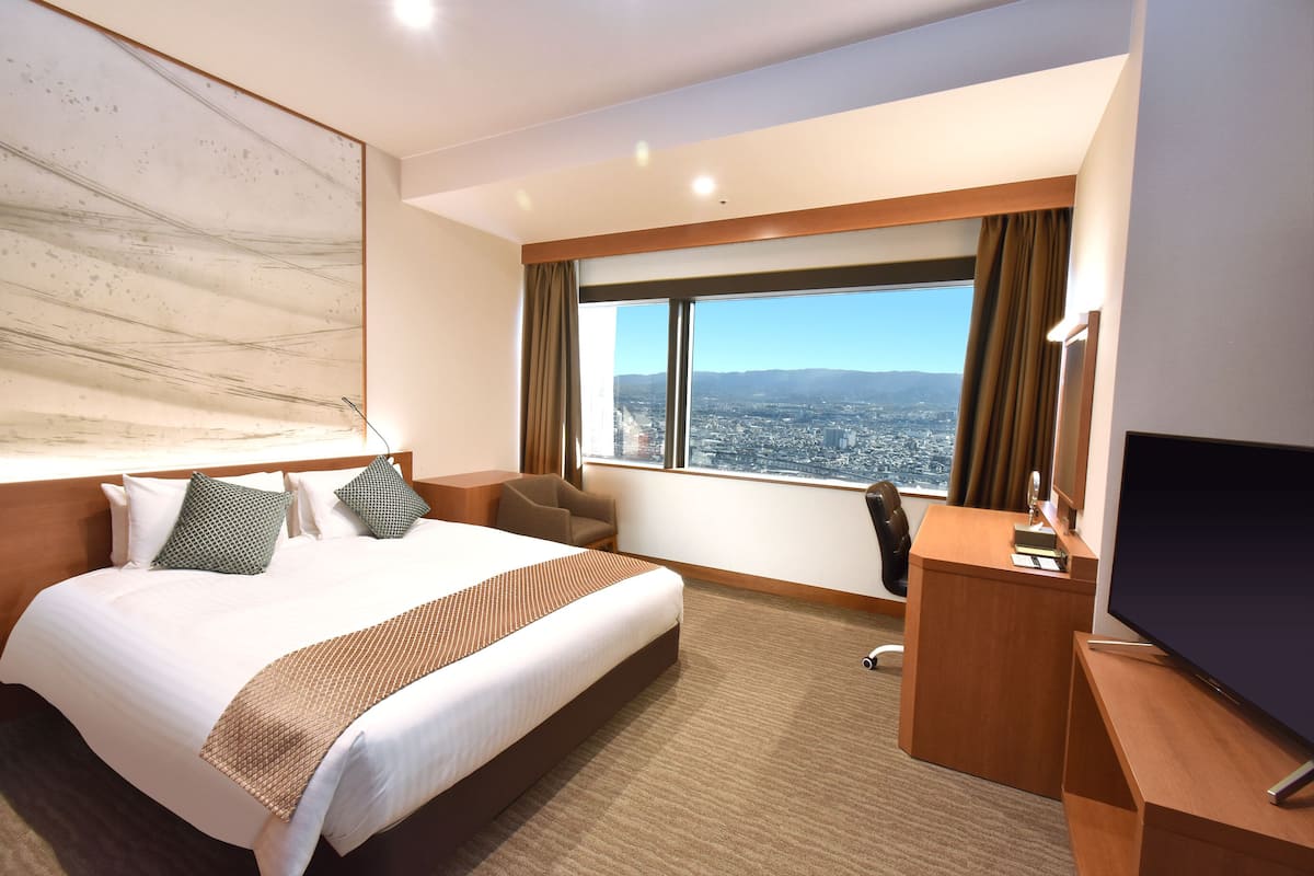 A hotel room with a large bed, a desk, a TV, and a view of a cityscape through a large window.