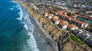 Vue aérienne d’un quartier résidentiel situé sur la côte, avec des bâtiments adossés à une falaise rocheuse et donnant sur l’océan.