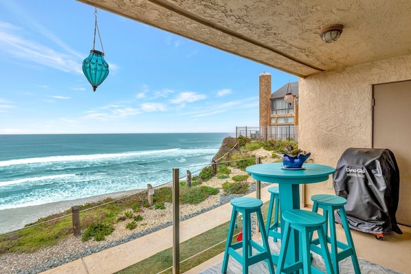 Un balcon avec vue sur l’océan, une table avec des tabourets, une lanterne suspendue et une housse de protection pour barbecue.