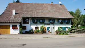 Kaksikerroksinen rakennus, jossa on tiilikatto, puuovi ja kukkaistutukset ikkunoiden alla.