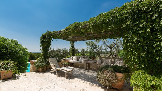 A patio area with a pergola covered in greenery, outdoor seating, and a pool in the background. - 339 I Trulli di Nanì by Perle di Puglia (Ostuni)