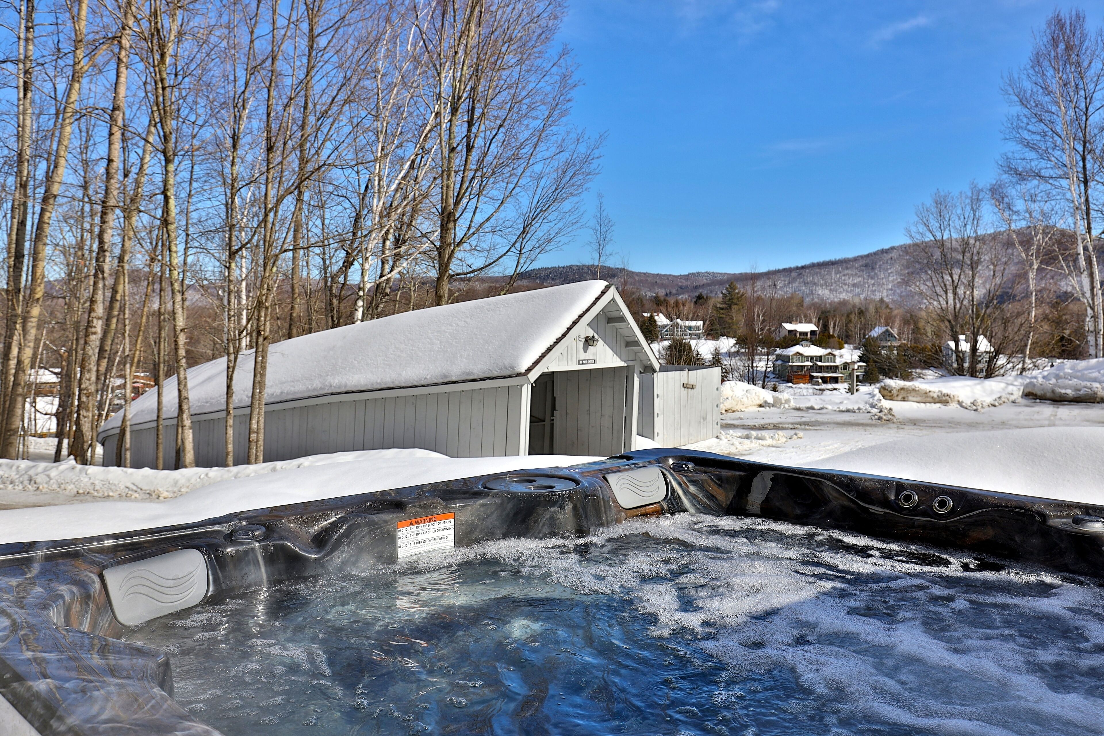 A hot tub with a wooden deck, surrounded by snow-covered ground and trees, with a building in the background.