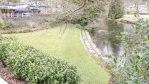 A riverside view with a stone pathway, grassy areas, and a building with a blue sign in the background. - Dolwen (Betws-Y-Coed)