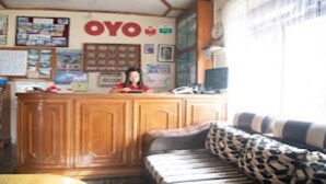 A reception area with a wooden desk, a sofa with patterned cushions, and a wall with various posters and a large 'OYO' sign. - MeroStay 015 (Pokhara)