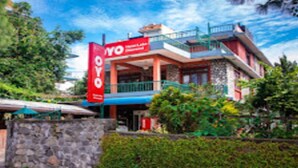 A hotel with a red and blue facade, a stone wall in front, and lush greenery around. - MeroStay 015 (Pokhara)