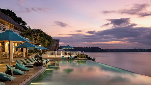 Una piscina infinita tranquila con sillones y sombrillas, con vista a una masa de agua calma al anochecer.