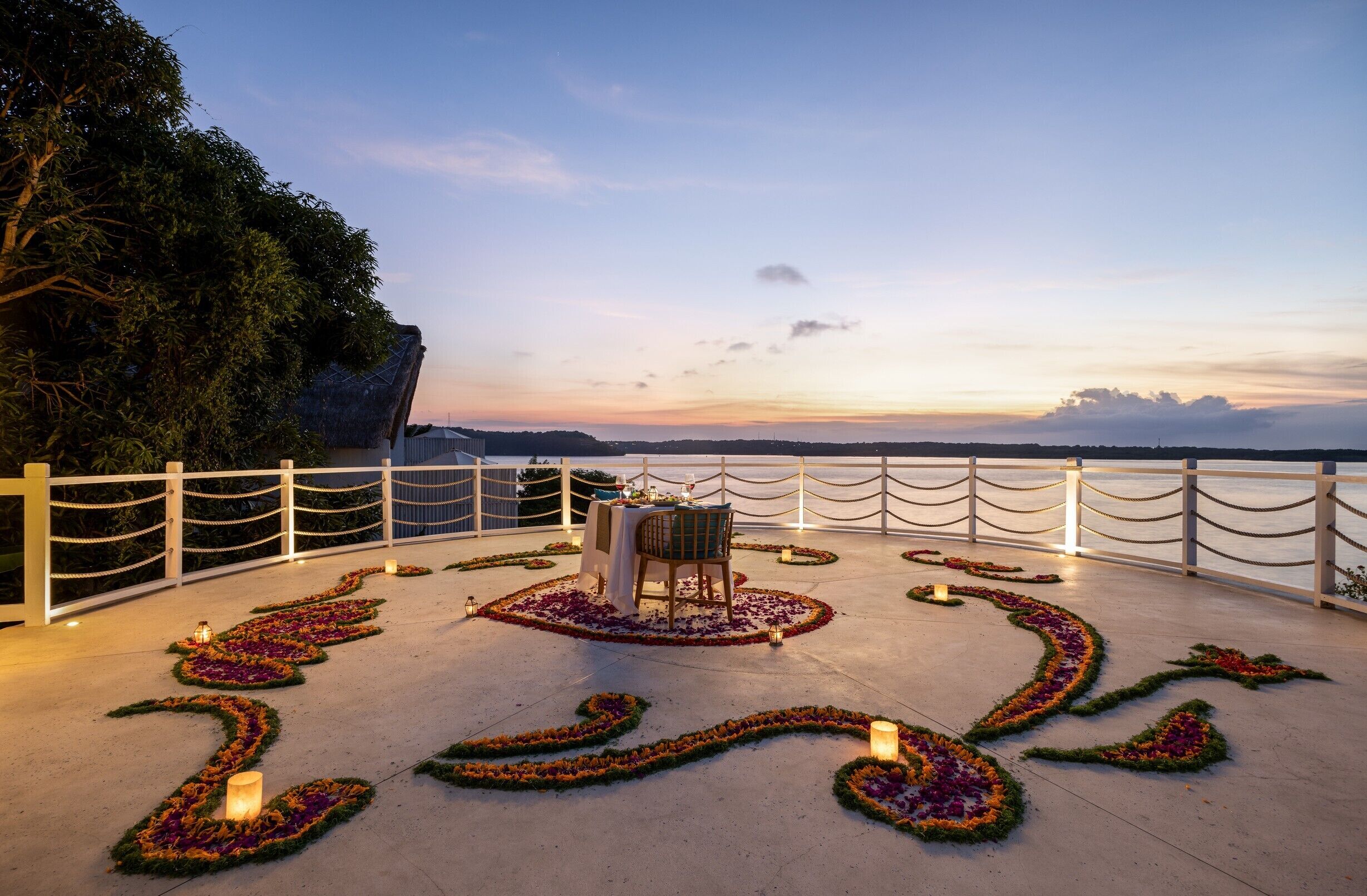 A sunset view from a balcony with a table set for two, surrounded by flower arrangements and a white fence.