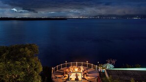 Terraza circular al aire libre con una chimenea central, rodeada por una barandilla, con vistas a una masa de agua y luces de la ciudad a lo lejos.
