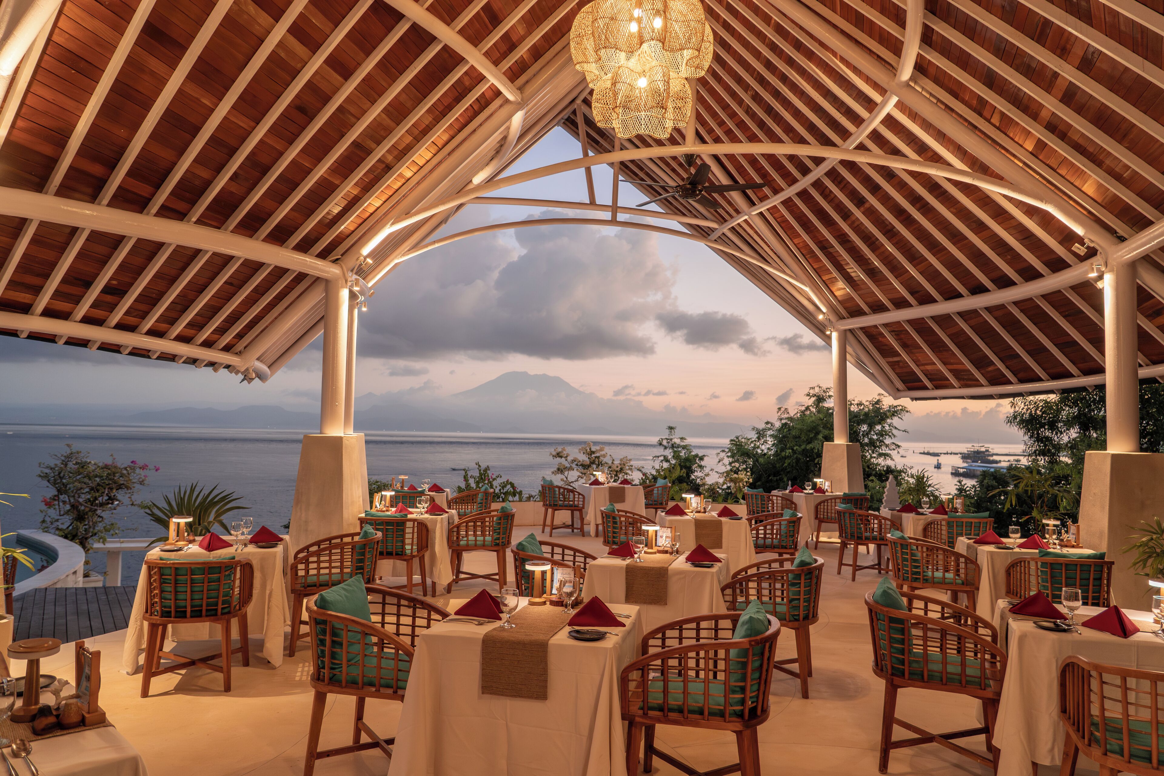 Un área de comedor al aire libre con mesas y sillas, vista al mar y un paisaje montañoso de fondo.