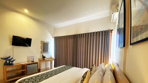 A hotel room with a bed, a desk with a computer, a TV mounted on the wall, and a window with curtains. - Mere Sea View Penida (Penida Island)