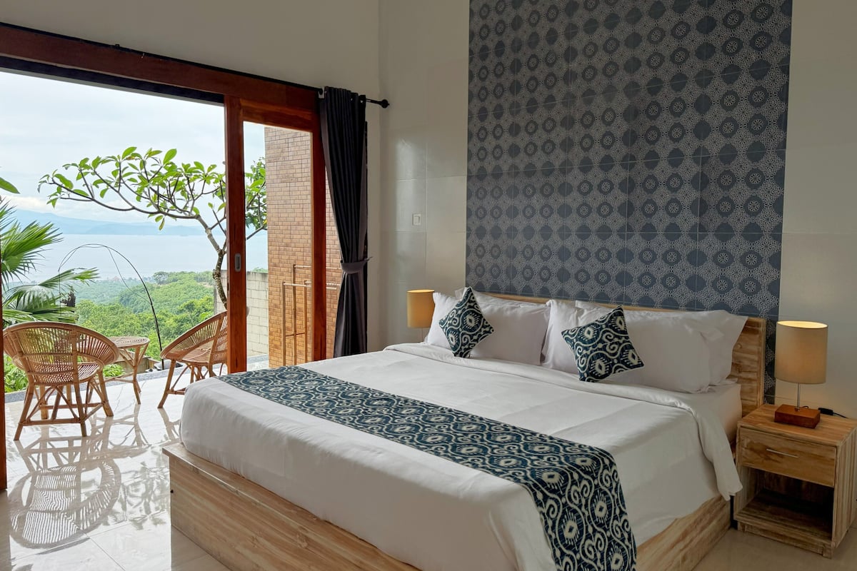 A bedroom with a large bed, two bedside tables, a view of greenery and a body of water through an open door.