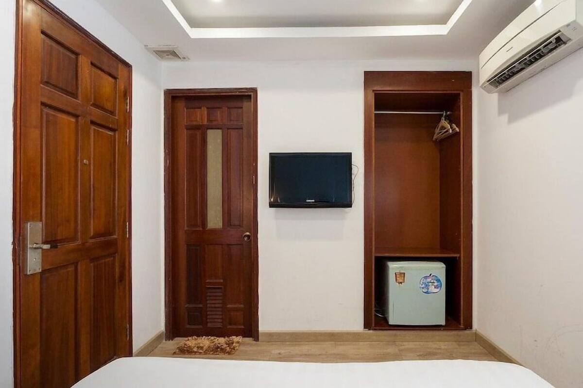 A bedroom with a wooden wardrobe, a television mounted on the wall, and a small refrigerator.