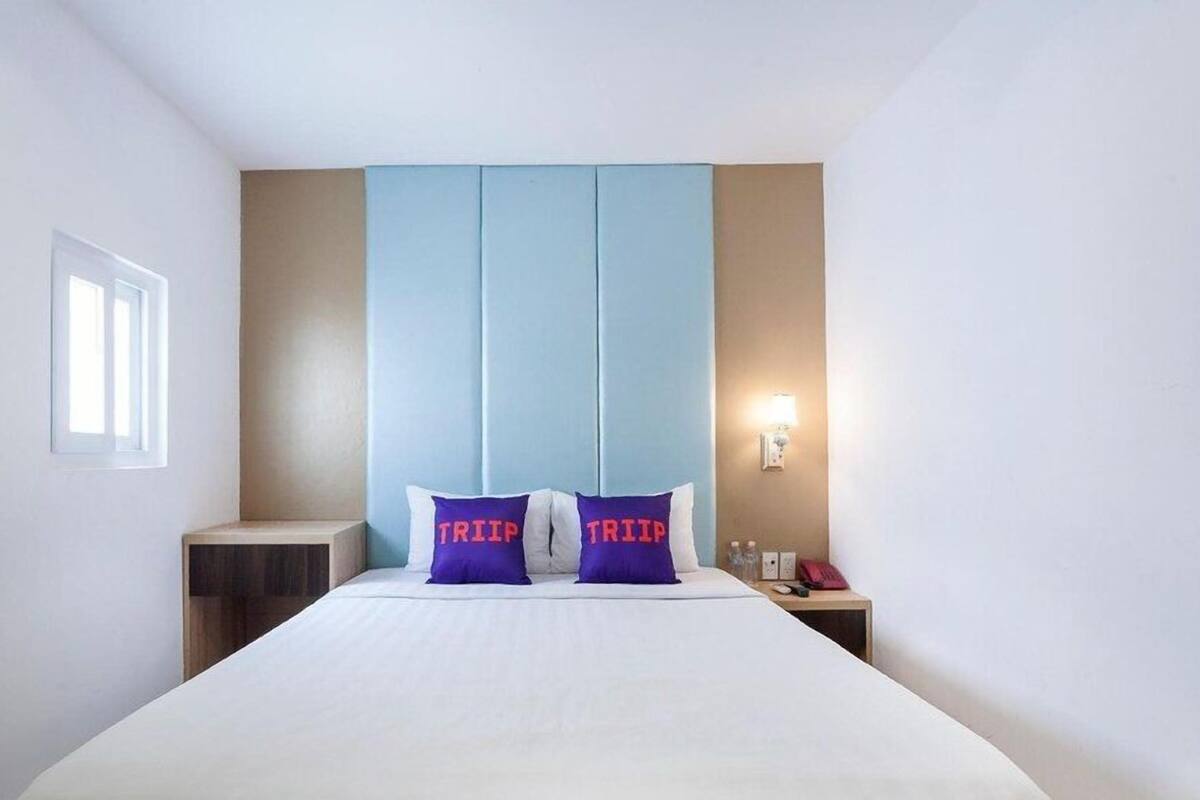 A hotel room with a bed, two pillows with 'TRIP' text, a nightstand, a lamp, and a window.