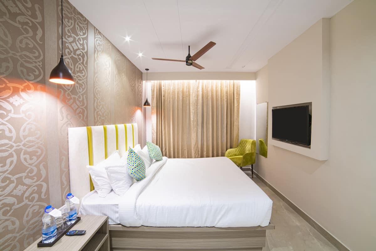 A hotel room with a bed, a TV, a ceiling fan, and a nightstand with a remote control and water bottles.