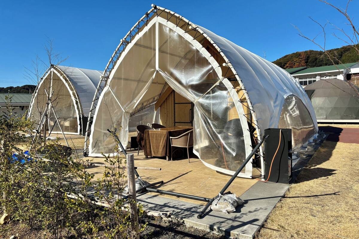 A series of dome-shaped tents with a transparent roof, equipped with a table and chairs inside, set on a paved area with a view of a forested hill in the background.